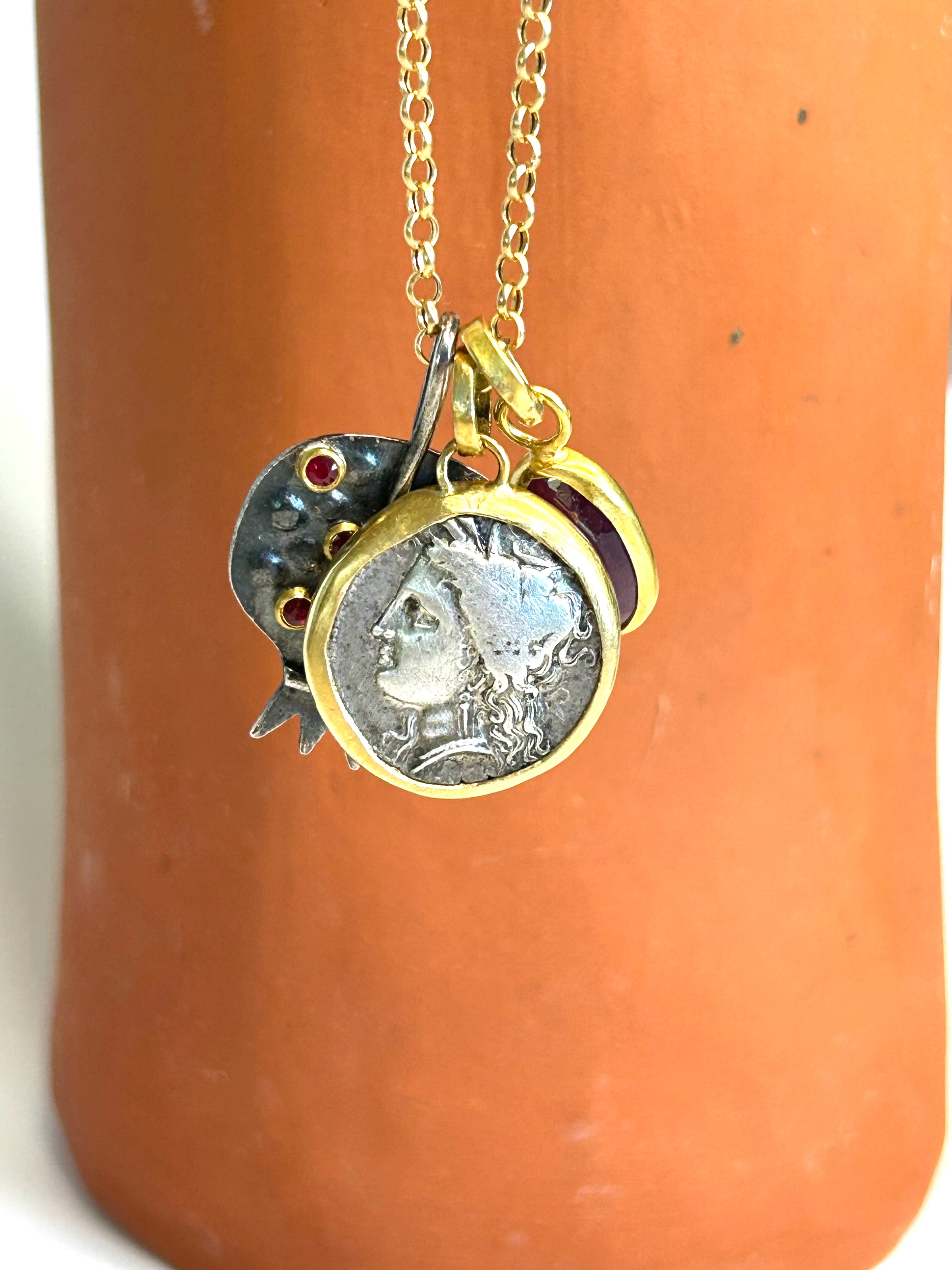 Gold chain necklace with a coin charm, a pomegranate pendant, and a ruby pendant on a terracotta surface